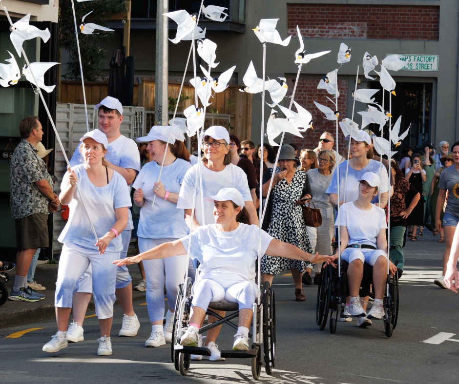 A group of WIDancers performing on the streets all wearing white while holding large white crafted birds on long poles high above their heads.